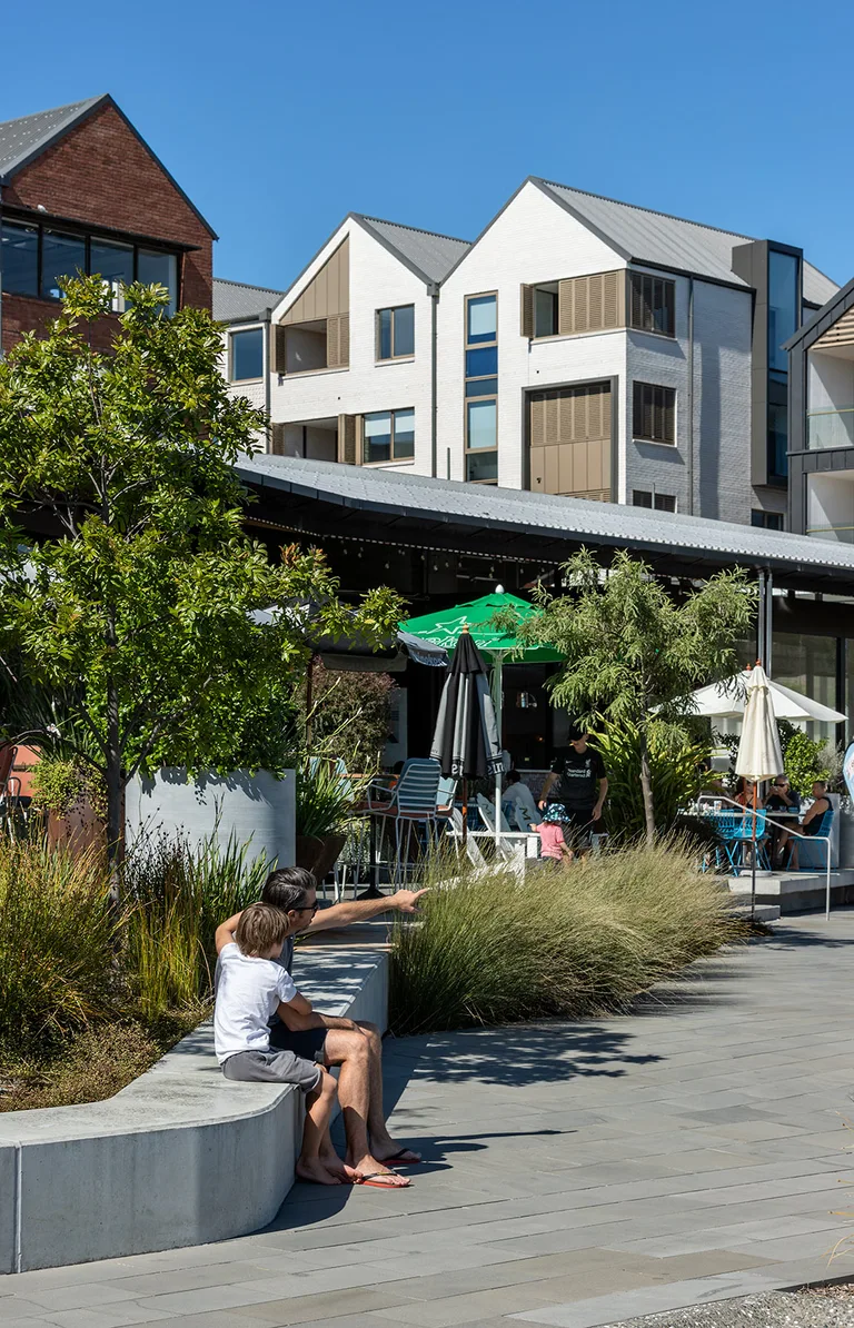 An adult and child seated outside the shops at Long Bay, looking and pointing to the right.